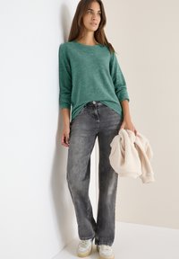 Teal gestreifter Pullover mit langen Ärmeln, graue weit geschnittene Jeans, hält eine hellbeige flauschige Jacke und weiße Sneakers mit Akzenten. Minimalistischer Hintergrund.
