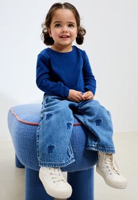 Plava košulja s dugim rukavima, plave traperice s uzorkom mašnica, bijele tenisice. Dijete sjedi na mekanom, zaobljenom plavom stolcu s narančastim obrubom.