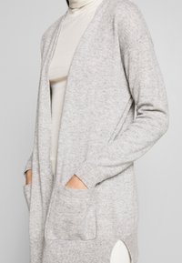 Ljust grå stickad cardigan med en lös passform, öppen framsida, långa ärmar och två framfickor. Sidoklyftor ger en subtil design Detalj.