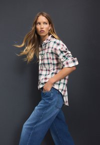 Camicia a quadri in tessuto morbido con tonalità rosse, verdi e bianche; abbinata a jeans blu comodi; vestibilità casual con maniche arrotolate.