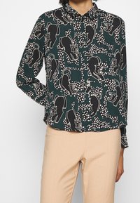 Groene button-up blouse met een zwart-wit berenpatroon en gestipte accenten. Lange mouwen, klassieke kraag, losse pasvorm en afgewerkte manchetten.