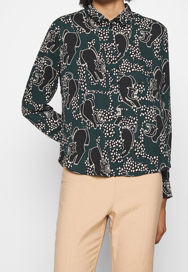 Vihreä nappipaitamalli, jossa on musta-valkoinen karhukuosi ja pilkulliset yksityiskohdat. Pitkät hihat, klassinen kaulus, rento leikkaus ja viimeistellyt hihansuut.