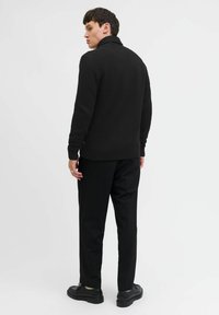 Schwarzer Strickpullover mit hohem Kragen, strukturierter Musterung, getragen mit schwarzen Hosen und schwarzen Schuhen, von hinten vor einem weißen Hintergrund gezeigt.