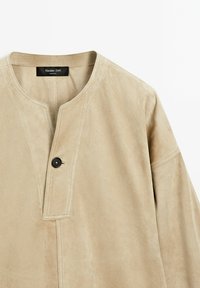 Beige Wildlederjacke mit einem runden Ausschnitt, Knopfverschluss und detaillierten Nähten entlang der Nähte. Einfaches, minimalistisches Design.