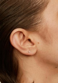 Pendiente de plata con una pequeña piedra brillante, en primer plano de una oreja con piel suave y cabello castaño liso.