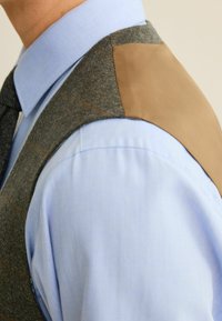 Grå tweedvest med brunt læder skulderlap, parret med en lyseblå skjorte og mørk slips. Har en skræddersyet pasform og subtilt ternet mønster.
