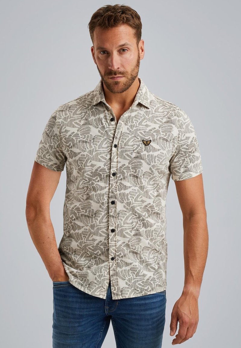 Kurzarm-Hemd mit Knopfleiste in hellem Beige und dunkelgrünem Blättermuster. Mit einem Logo auf der Brust und schwarzen Knöpfen, kombiniert mit blauen Jeans.