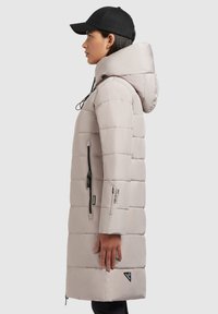 Manteau rembourré de couleur beige clair, avec un col montant, une capuche et des poches zippées noires. Présente un accent de logo sur le bas de l'avant et un tissu texturé.