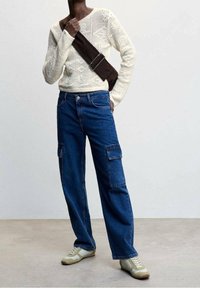 Lichtcrème gebreid sweater, blauwe losse cargo jeans met zijzakken en beige sneakers, allemaal tegen een neutrale achtergrond.