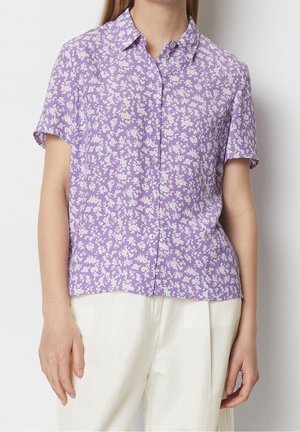 Femme portant un chemisier boutonné à manches courtes avec un motif floral blanc sur tissu lavande et un pantalon blanc taille haute.