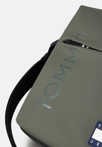 Grüne wasserdichte Tasche mit glänzendem "TOMMY"-Logo. Verfügt über eine glatte Oberfläche, einen schwarzen Tragegurt und ein marineblaues Akzent mit einem Etikett-Detail.