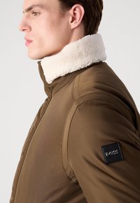 Brun pufferjakke med en cremefarvet shearlingkrave, der har et sort BOSS-logo patch på venstre skulder og glat, tekstureret stof.
