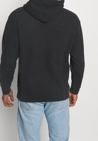 Sweat à capuche noir à manches longues, avec poignets côtelés et poche kangourou. Porté avec un jean denim bleu clair.