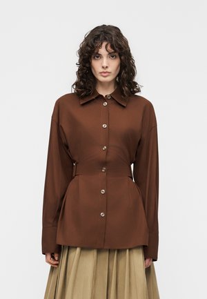 Vrouw met schouderlang krullend haar, gekleed in een bruin, met knopen gesloten, top met lange mouwen en een beige geplooide rok tegen een witte achtergrond.