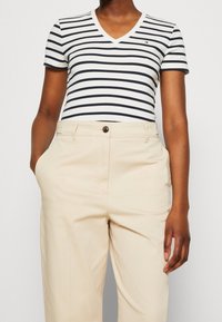 Pruhované tričko s V-výstřihem v bílé barvě s námořnickými pruhy, spárované s béžovými kalhotami na míru. Tričko má krátké rukávy a akcent v podobě loga.