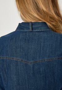 Tamsiai mėlynas džinsinis švarkas su smailiu atsegamu nugaros siūlu, matomomis siuvimo detalėmis ir smailia apykakle. Tekstūra atrodo lygi ir šiek tiek dėvėta.