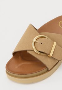 Sandale à talon en suède beige avec une large lanière ornée d'une boucle circulaire en ton doré et une semelle intérieure marron clair sur une semelle épaisse et rembourrée.