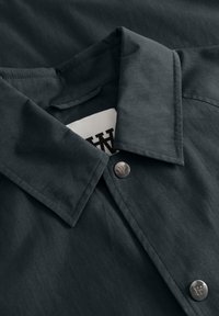 Tumesinine nööbitav särk terava kraega, millel on metallnööbid, millele on graveeritud "W" logo, ja osaliselt nähtav valge silt sees.