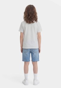 Παιδί με μεσαίου μήκους κατσαρά μαλλιά, φοράει απλό λευκό μπλουζάκι, ανοιχτό μπλε τζιν σορτς, λευκές κάλτσες και λευκά αθλητικά παπούτσια, στέκεται με την πλάτη προς τα έξω.