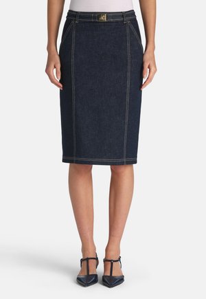 Jupe crayon en denim foncé avec garniture dorée à la taille, détails de coutures apparentes et silhouette ajustée. Associée à des chaussures pointues.