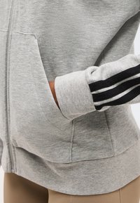 Persona, kas valkā gaiši pelēku rāvējslēdzēja džemperi ar melnām un baltām svītrām uz piedurknes, roka ievietota priekšējā kabatā, saskaņota ar bēšām biksēm.