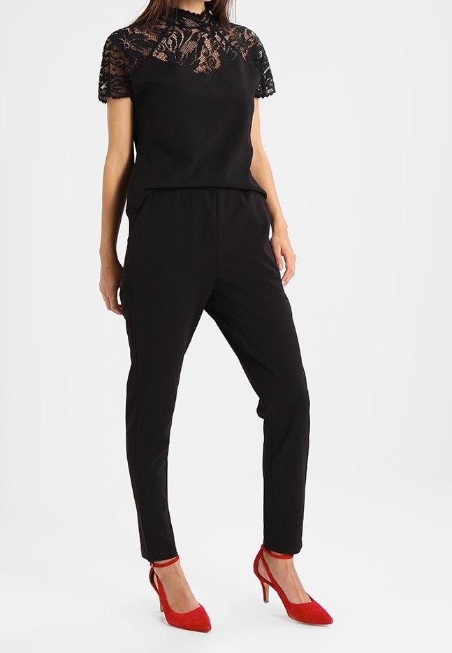 Frau trägt ein schwarzes Spitzenoberteil, schwarze schmale Hose und rote High-Heel-Schuhe mit Knöchelriemen, steht vor einem schlichten Hintergrund.