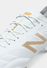 Chuteira de futebol branca com um upper texturizado, apresentando detalhes em dourado, uma língua em malha e atacadores. Logótipo "N" notável com riscas diagonais de lado.
