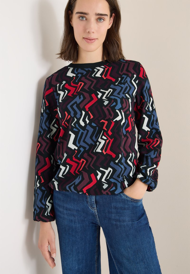 Langærmet top med et flerfarvet zigzag-mønster i rød, hvid, blå og sort, parret med blå denimjeans. Blød tekstur.