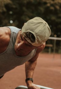 Sportos férfi szürke ujjatlan pólóban fekvőtámaszokat végez, világos színű perforált sapkát és fekete karórát visel, piros felületen.