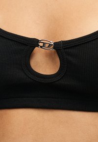 Gros plan d'un crop top noir côtelé avec une petite ouverture en forme de trou de serrure fermée par un fermoir ovale argenté au niveau du décolleté.