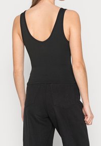 Svart ribbad tanktop med djup V-nacke, figurnära design och breda axelband. Tyget har en strukturerad yta. Kombinerad med svarta högmidjade byxor.