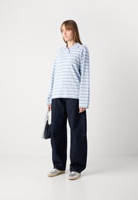 Camicia azzurro chiaro a maniche lunghe con righe orizzontali, abbinata a jeans scuri e sneakers trasparenti. Il modello tiene una borsa trasparente.