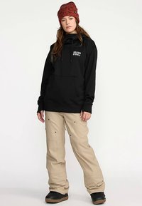 Svart hútröja med framficka och vitt logotyp. Beige snöbyxor med flera fickor och svarta kängor. Brun mönstrad mössa.