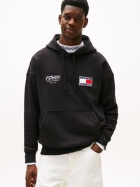 Sudadera negra con bolsillo frontal, que presenta un logo bordado en blanco y detalles rectangulares en rojo, blanco y azul en el pecho. Combinada con pantalones claros.
