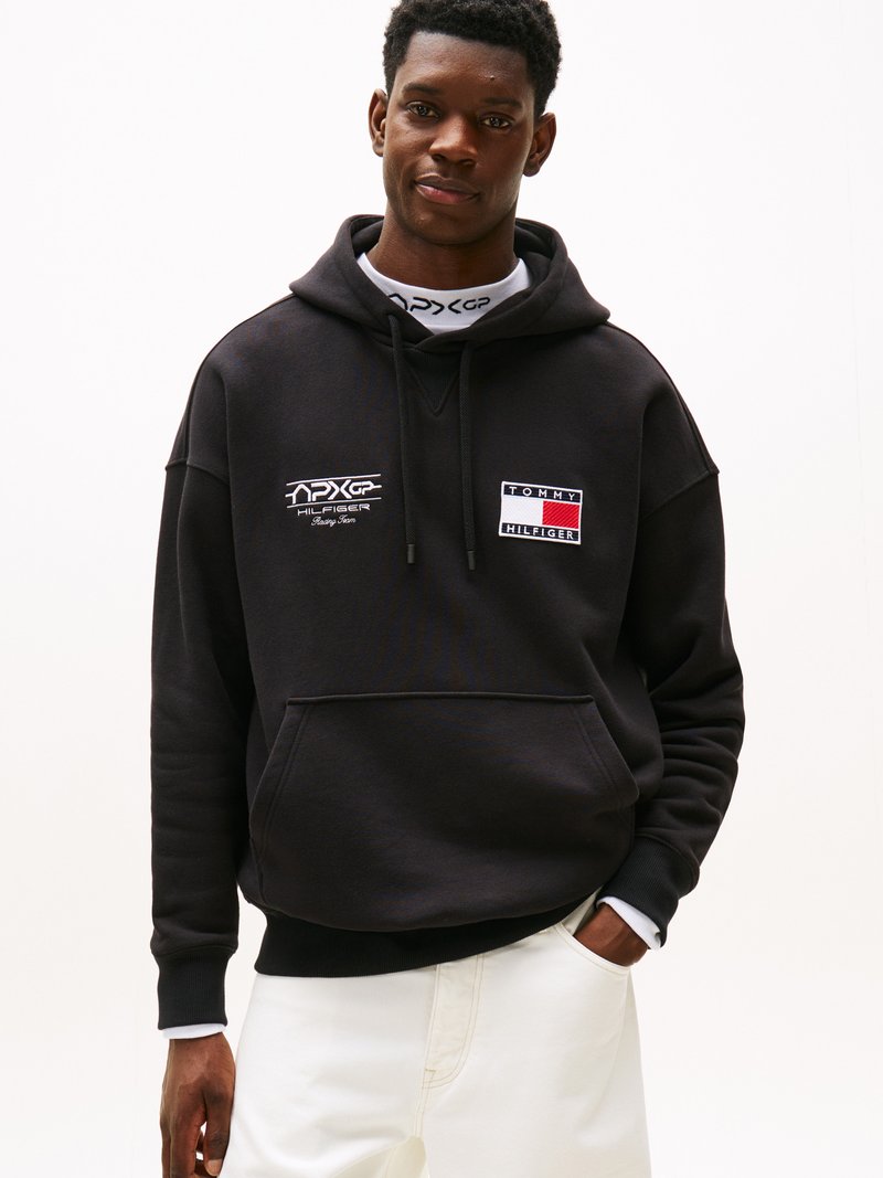 Sudadera negra con bolsillo frontal, que presenta un logo bordado en blanco y detalles rectangulares en rojo, blanco y azul en el pecho. Combinada con pantalones claros.