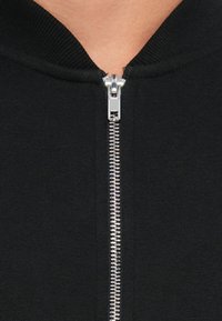 Sort lynlås hoodie med sølvmetal lynlås, ribbet krave og glat tekstur, med syninger langs lynlås linjen.