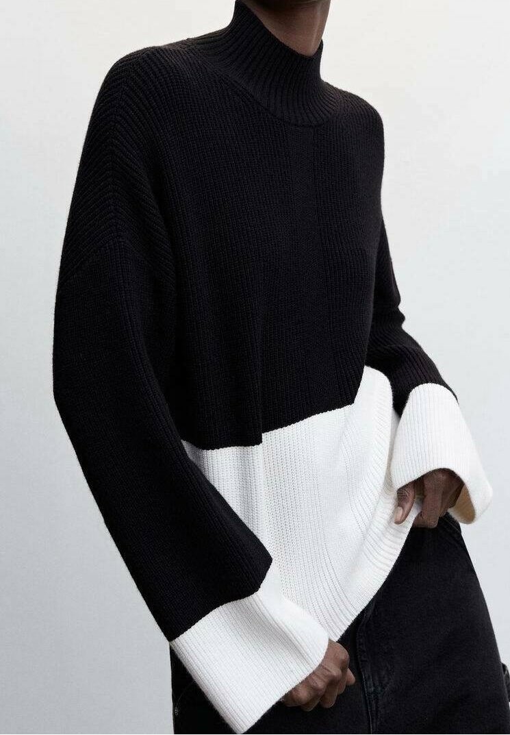 Personne portant un pull à col roulé à blocs de couleur noir et blanc côtelé avec des manches longues, les mains détendues sur les hanches, sur un fond uni.