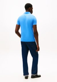 Lyseblå poloshirt med korte ermer, glatt tekstur og krage; kombinert med mørkeblå jeans og svarte sko, sett bakfra.