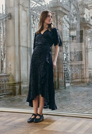 Kobieta w ciemnej kwiatowej sukience midi z kopertowym dekoltem i czarnych balerinach stoi w pomieszczeniu blisko dużego szklanego okna z ozdobną kutą kratą i widoczną elewacją kamienną.