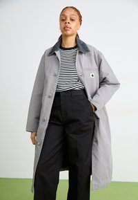 Cappotto grigio lungo con un collo scuro, indossato sopra una camicia a righe bianco e nera, abbinato a pantaloni neri. Design semplice, senza motivi visibili.