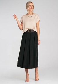 Beige kurzärmliges Oberteil, kombiniert mit einem schwarzen Faltenrock, der in der Taille mit einem braunen Gürtel gerafft ist und mit einer zarten Halskette accessoriert wird.