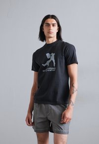 Sort bomulds t-shirt med grafisk tryk af en person, der bærer en taske. Parret med grå shorts. Modellen står foran en neutral baggrund.