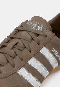 Καφέ σουέντ αθλητικό παπούτσι με γκρι κορδόνια, που διαθέτει τρεις ασημένιες ρίγες, υφή στην επιφάνεια και διακοσμητική γλώσσα από δέρμα με σήμανση της μάρκας.