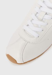 Λευκό δερμάτινο αθλητικό παπούτσι με λευκά κορδόνια και καφέ καουτσούκ σόλα, που δείχνει λεπτομερή ραφή στην περιοχή του δακτύλου.