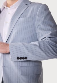Helles blaues Blazer mit Streifen aus strukturiertem Stoff, ausgestattet mit einer einzelnen Tasche und vier schwarzen Knöpfen am Ärmelsaum.