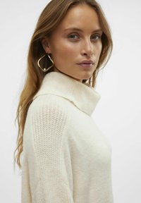 Maglione color crema con colletto alto, con maniche a maglia testurizzata e corpo liscio. Grandi orecchini a cerchio dorati con dettaglio di perla.