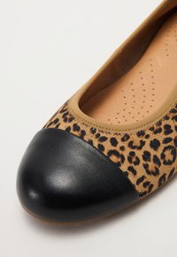 Ballettschuhe mit Leopardenmuster, schwarzer Kunstleder-Spitze und braunem Elastikband. Das Innenfutter ist beige und hat runde Perforationen zur Luftzirkulation.