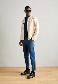 Beige dzseki két elülső zsebbel, tengerészkék póló alatta, kék nadrág és fekete fűzős cipő. Simább textúra, modern szabás.