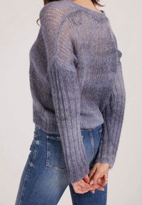 Personne portant un pull ample semi-transparent violet clair à poils longs avec des manches longues, associé à un jean en denim bleu, les mains jointes derrière le dos.