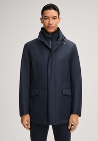 Marineblauwe jas met een staande kraag, een rits aan de voorkant en twee grote zijzakken. Glad, gestructureerd stof met een getailleerde pasvorm.